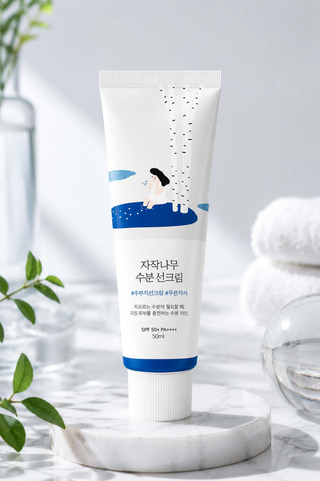 ROUND LAB Birch Juice Moisturizing Sunscreen SPF50+ PA++++ 50ml elegantly displayed on a white marble tray in a bright minimalist skincare setting with soft natural light, fresh green botanical accents, clear glass décor and folded white towels, highlighting its hydrating birch sap formula, lightweight texture, daily sun protection and clean Korean skincare aesthetic for healthy, radiant skin.